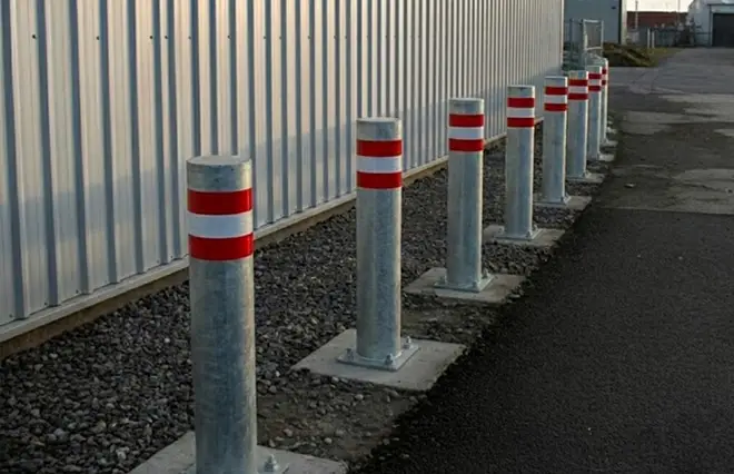 Fencing featuring a row of galvanized steel bollards with red and white reflective bands alongside an industrial building.